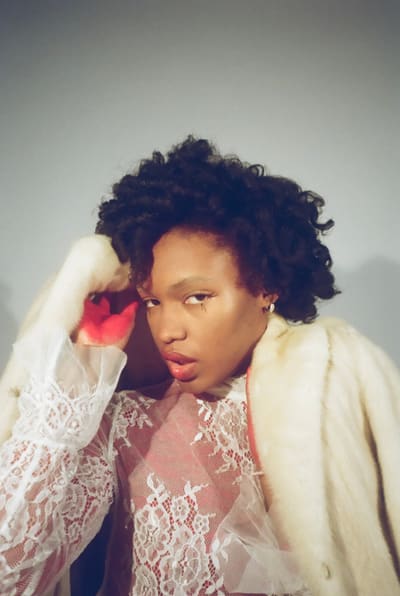 Person with curly hair in a lace top and cream fur coat, red-stained hand raised to the head