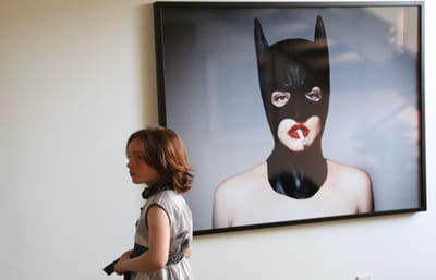 Young model in a grey dress standing beside a large framed photo of a figure in a black hood