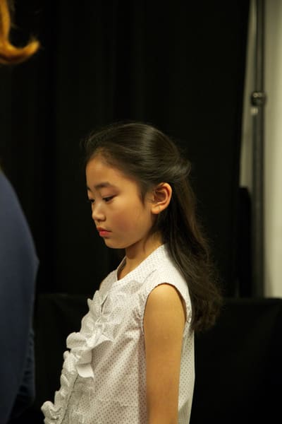 Young model in a white ruffled top looking down against a black curtain