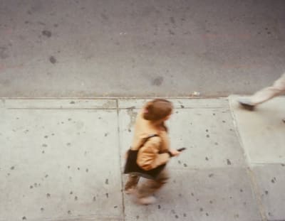 Motion-blurred person in a tan jacket walking along a gum-spotted sidewalk, seen from above