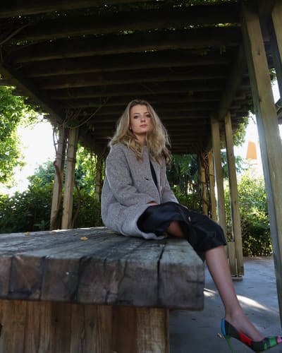 Model in a grey coat seated on a rough wooden table under a pergola