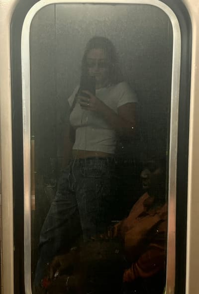 Selfie reflected in a subway car door window: person in white T-shirt and jeans holding up a phone