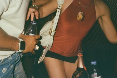 Torso of a person in a rust halter top with a white shoulder bag, champagne bottle in front