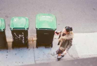 Person in a fur-trimmed parka walking past green-lidded trash bins, seen from above
