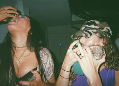 Two people drinking side by side, one tipping back a bottle and one in a camouflage cap